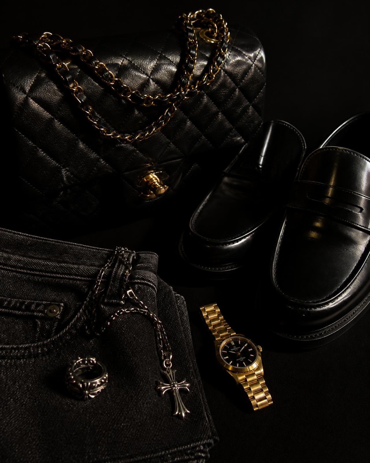 Editorial dark flat-lay of a quilted leather handbag, leather loafers, a gold watch, silver jewelry and black denim